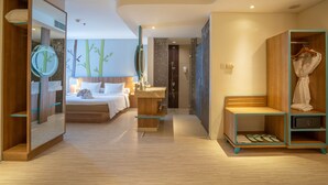A modern hotel room with a wooden floor, a bed with white linens, a bedside table, a mirror, a wardrobe, and a shower area. - Grand Livio Kuta Hotel (Legian)