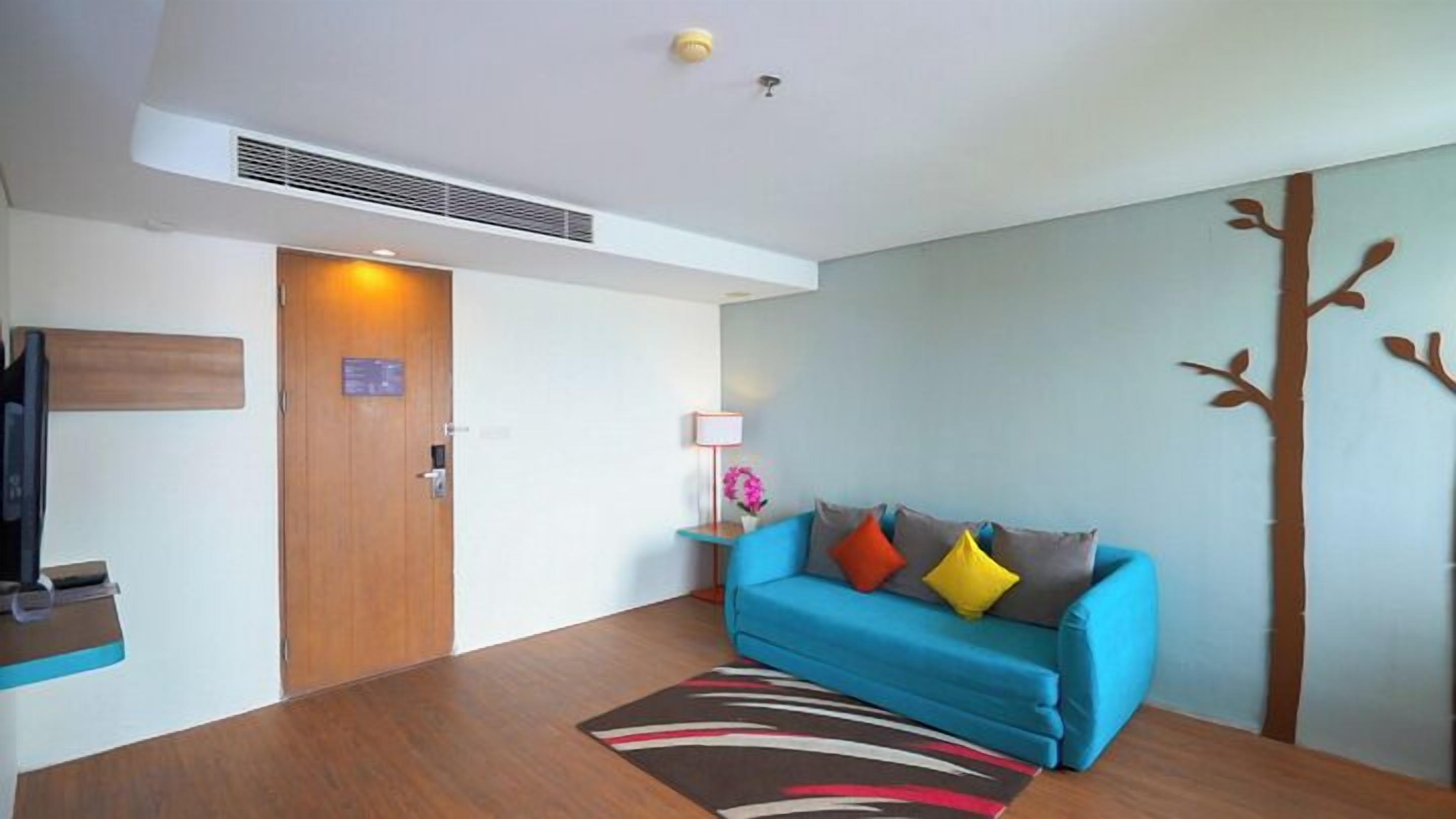 Una habitación de hotel moderna con una puerta de madera, un sofá azul con cojines de colores, una mesita auxiliar con lámpara y un equipo de aire acondicionado instalado en la pared.
