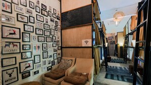 A cozy room with a wall adorned with framed artwork, a comfortable sofa with cushions, and a small woven stool. - Wire Hostel Patong (Patong)