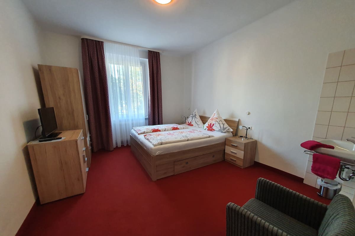 A bedroom with a bed, a wooden dresser, a desk, a chair, a bedside table, a lamp, and a bathroom with a sink and a mirror.