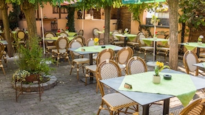 Un café en plein air avec des tables et des chaises en osier, entouré d’arbres et de plantes en pot.
