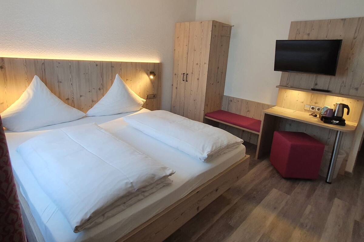 A hotel room with a wooden bed, a desk with a flat-screen TV, and a red bench.