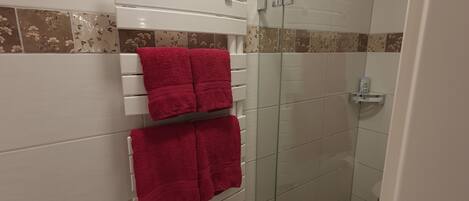 A bathroom with white tiles, a glass shower enclosure, and a towel rack with red towels.