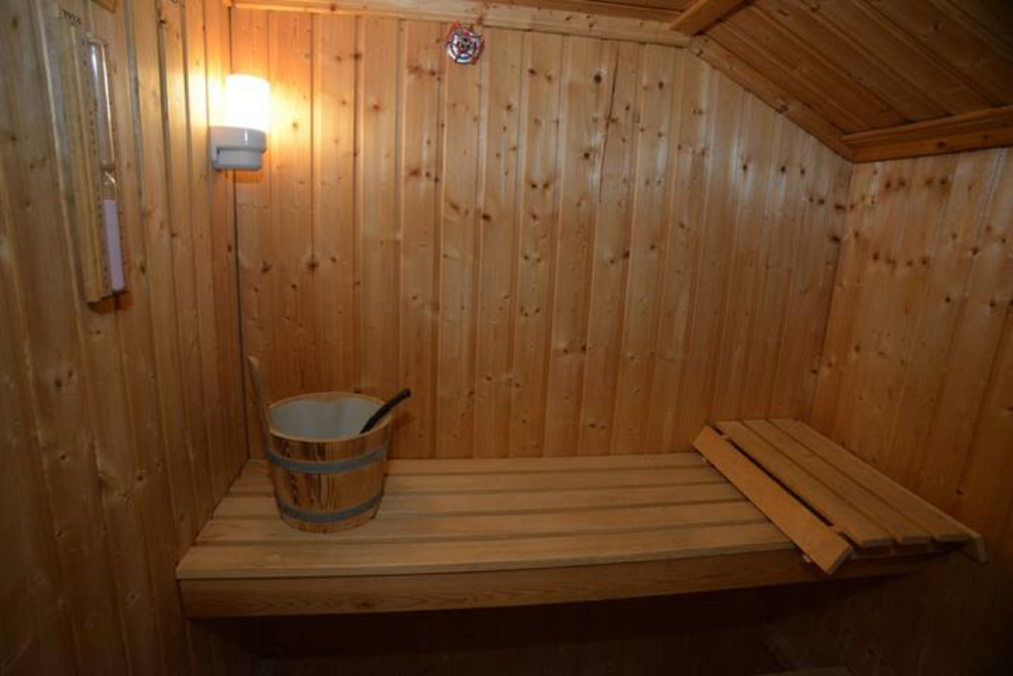 A wooden sauna with a bench, a bucket, and a wall-mounted light.