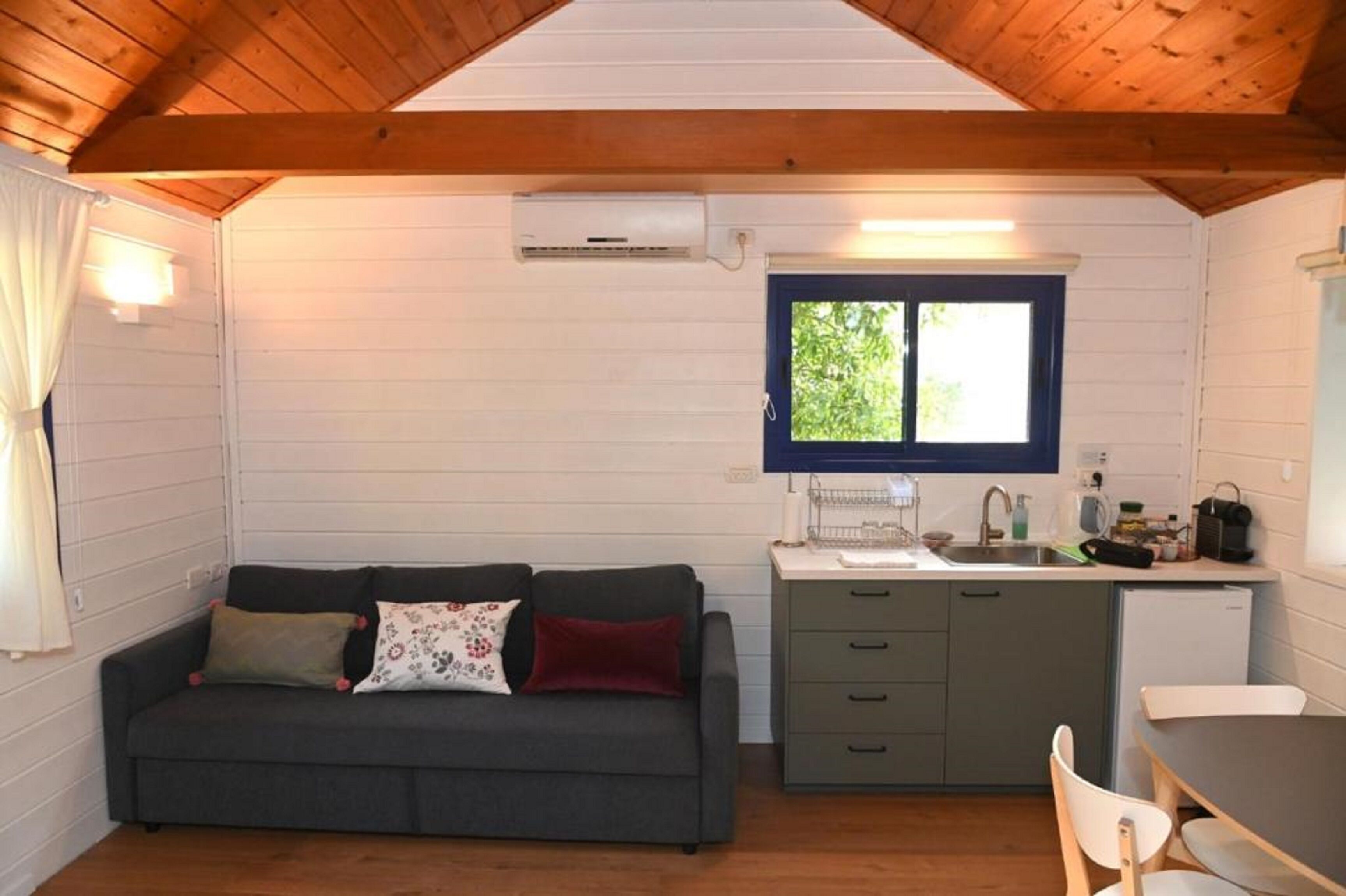 Een knus ingerichte kamer met een grijze bank, een klein keukentje en een raam met uitzicht op groen.