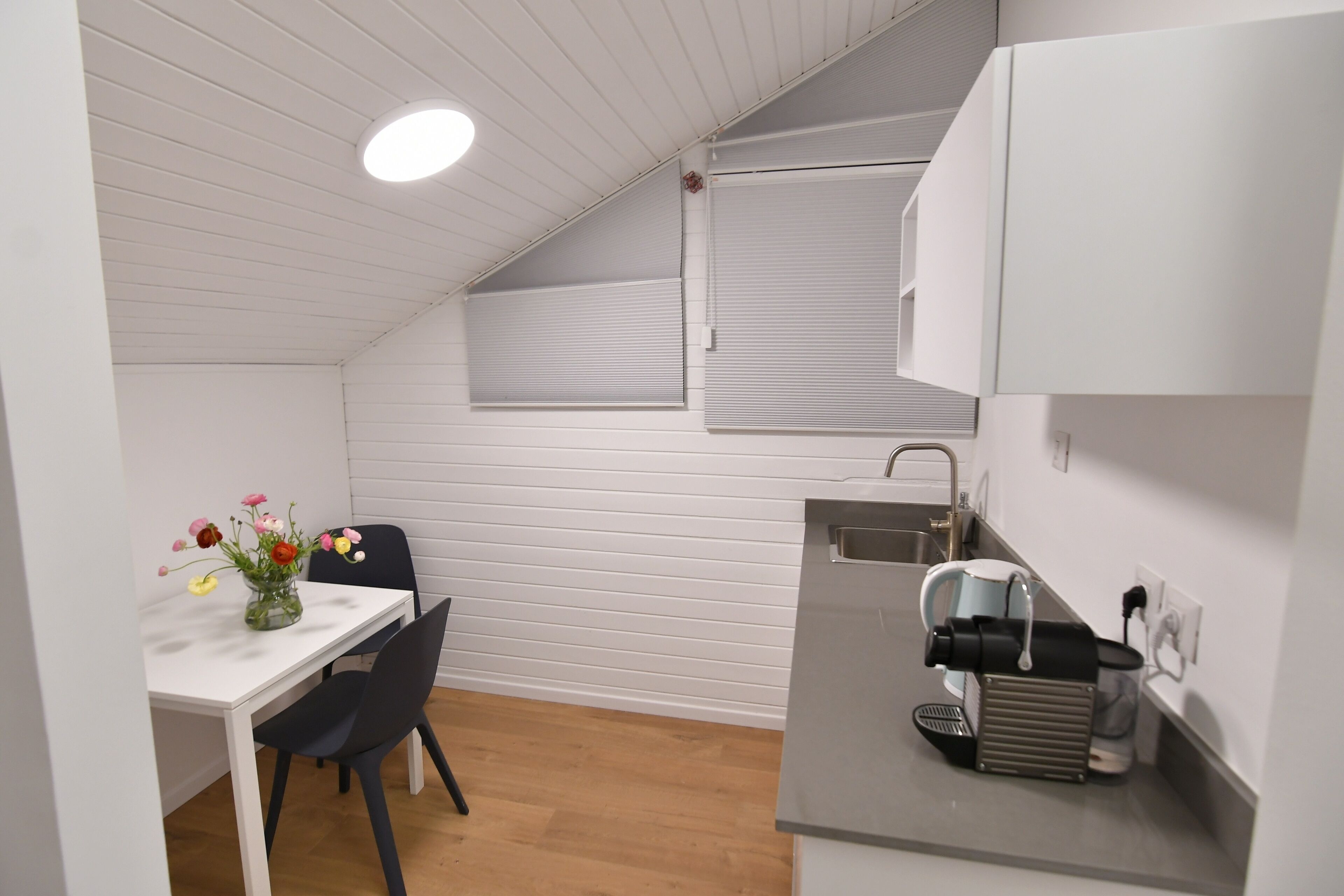 Una kitchenette pequeña con encimera blanca, un lavamanos y una cafetera. Hay una mesa de comedor con un florero y una silla.