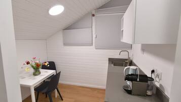 Una kitchenette pequeña con encimera blanca, un lavamanos y una cafetera. Hay una mesa de comedor con un florero y una silla.