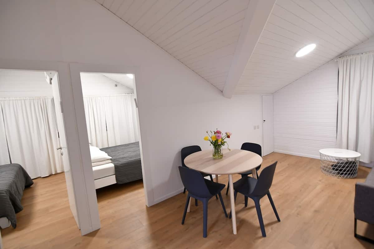 Ein modernes, gut beleuchtetes Zimmer mit Holzboden, einem runden Tisch mit Stühlen, einem Bett mit grauem Kopfteil und einer weißen Decke mit Einbauleuchten.