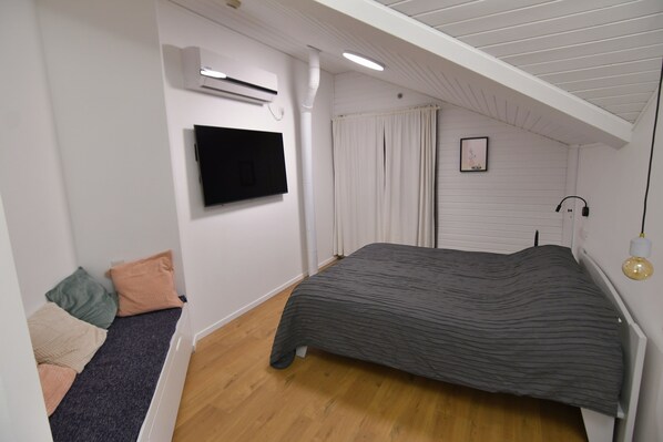 Une chambre à coucher comprenant un lit, un téléviseur fixé au mur, un appareil de climatisation et une fenêtre avec des rideaux.