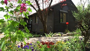 Un chalet rustique en rondins de bois, entouré d’un jardin où poussent différentes sortes de fleurs et d’un arbre aux feuilles rouges.