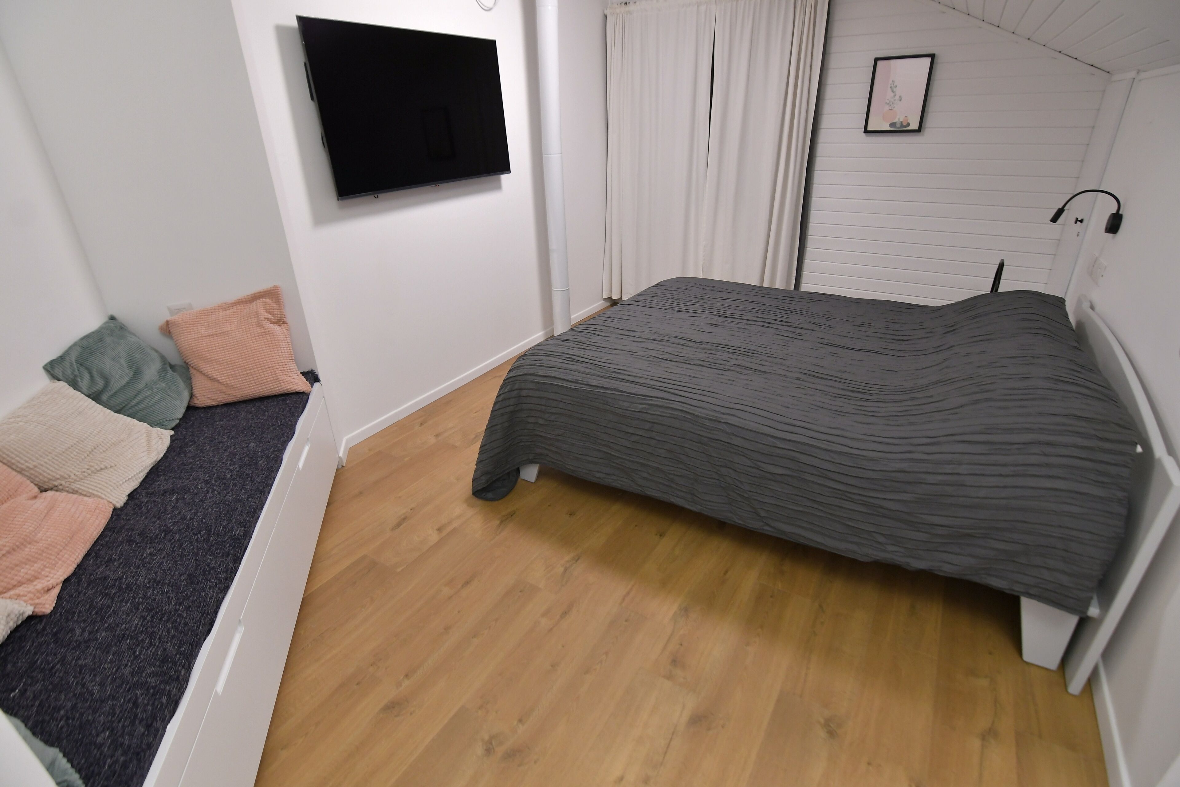 Un dormitorio con cama, televisor montado en la pared, ventana con cortinas y mesita de noche con lámpara.