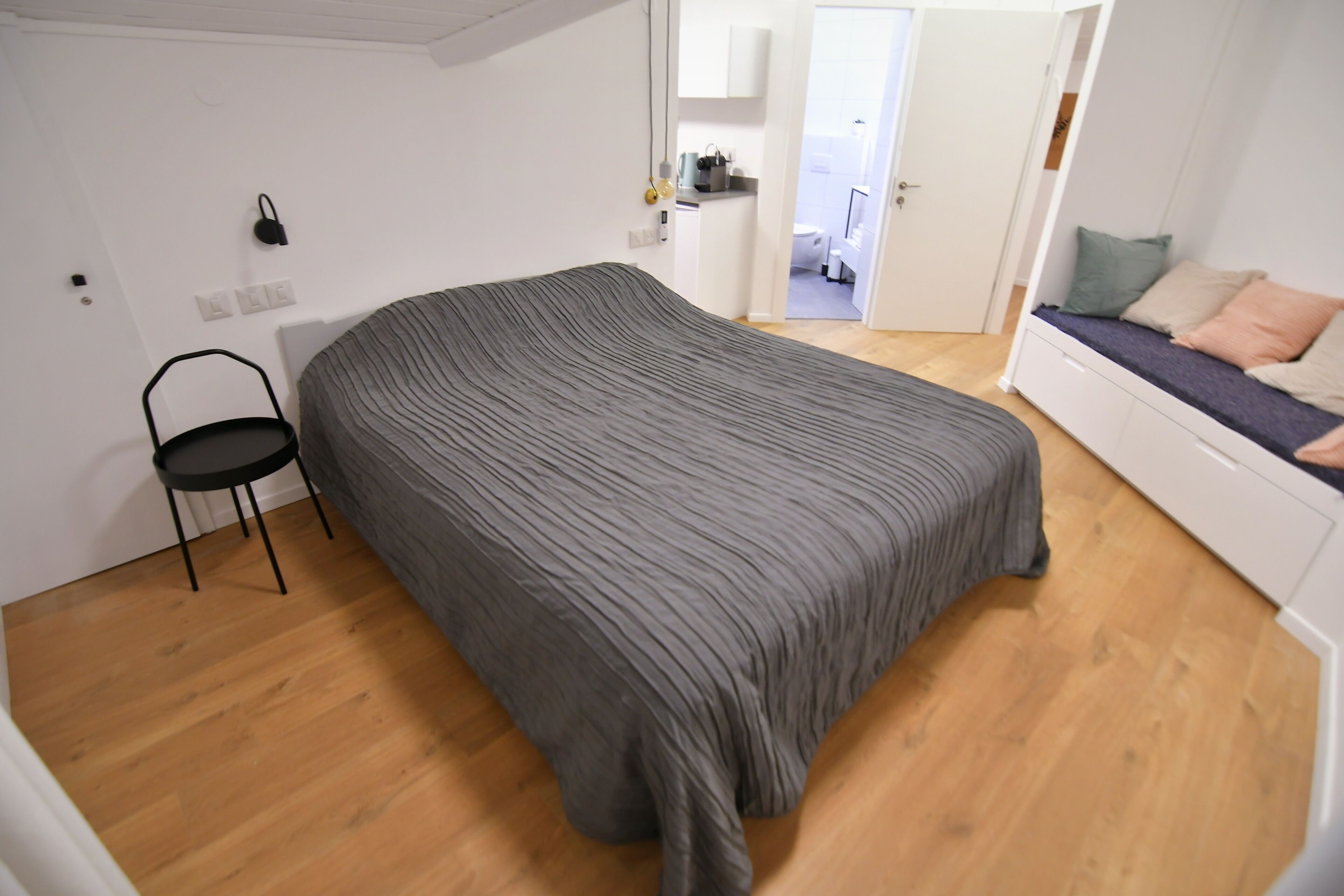 Una habitación con una cama grande, una silla, una kitchenette y un baño visible a través de una puerta abierta.