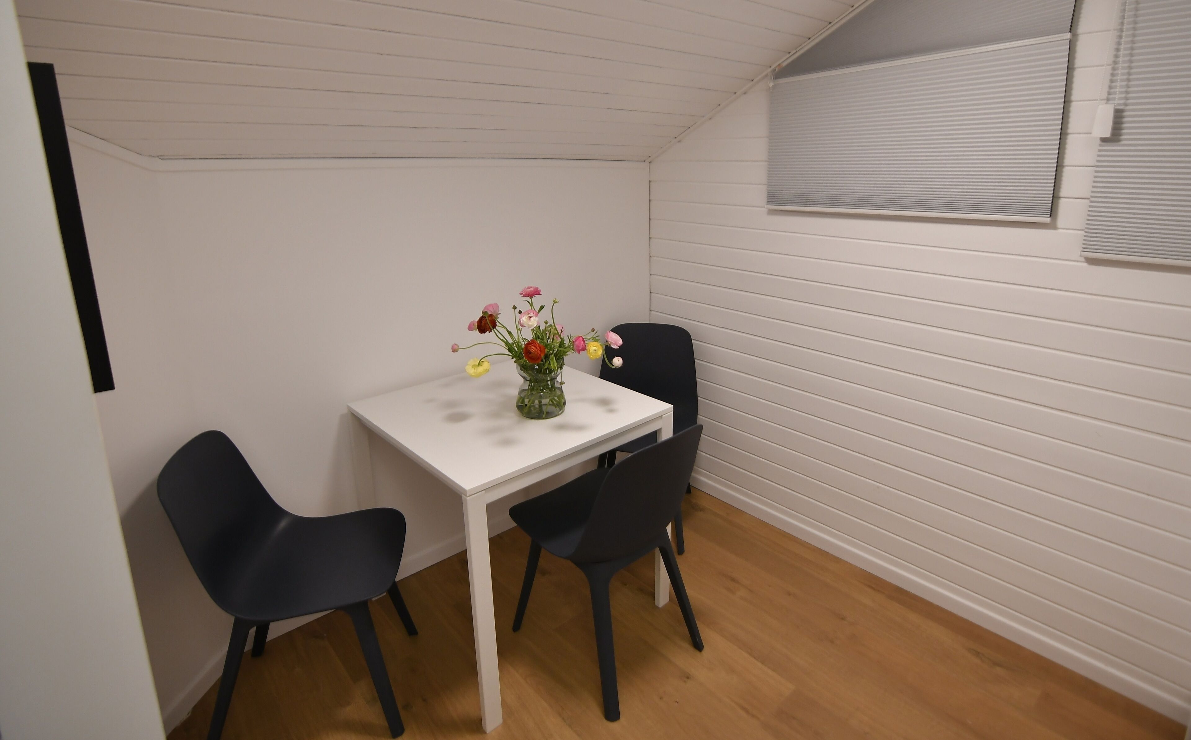 Een gezellig eethoekje met een witte tafel en twee zwarte stoelen, een vaas met bloemen, en een raam met zonwering.