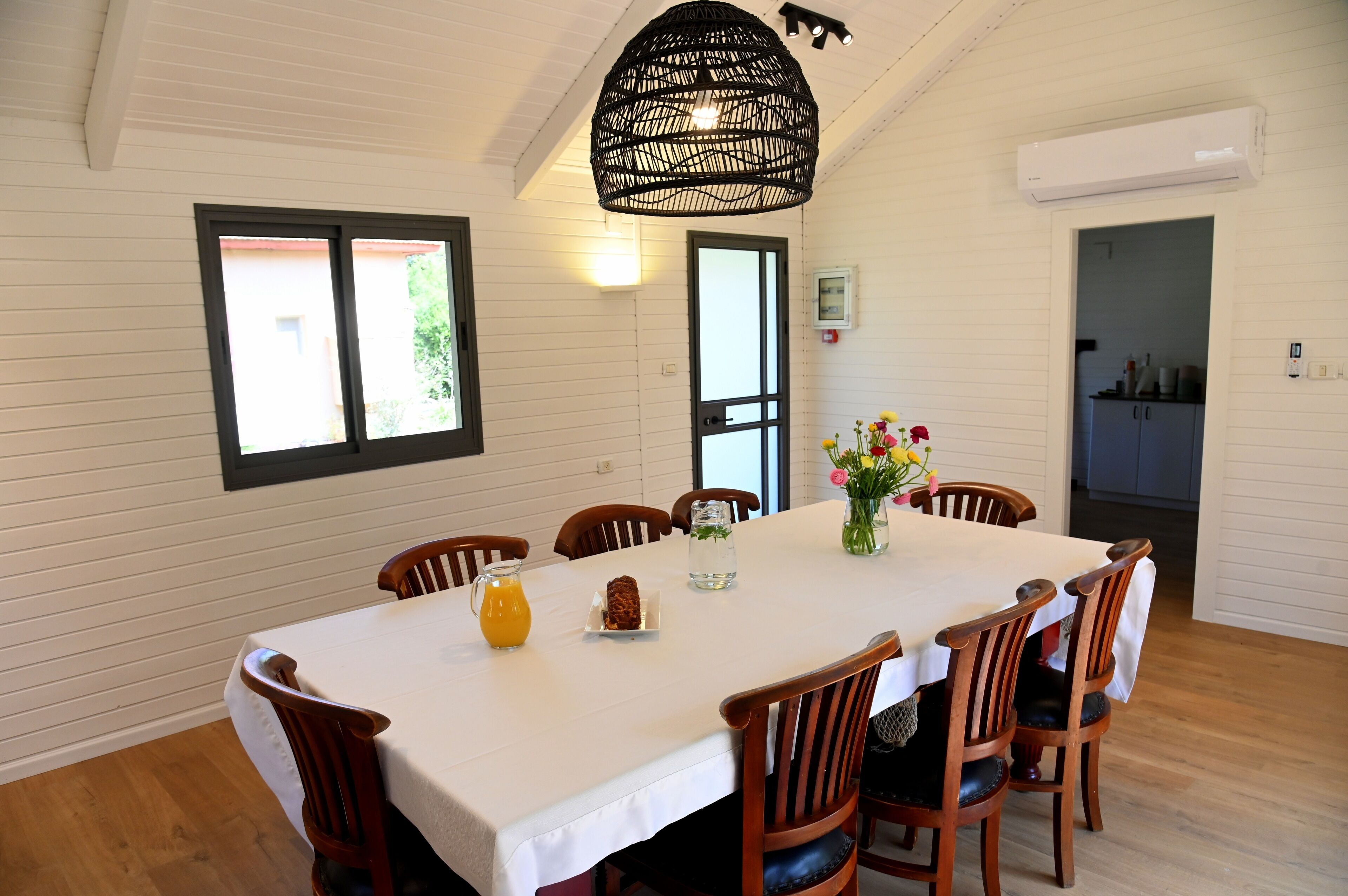 Un coin repas avec une table blanche, des chaises en bois, un luminaire noir suspendu et une fenêtre avec des stores.