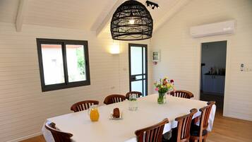 Un coin repas avec une table blanche, des chaises en bois, un luminaire noir suspendu et une fenêtre avec des stores.