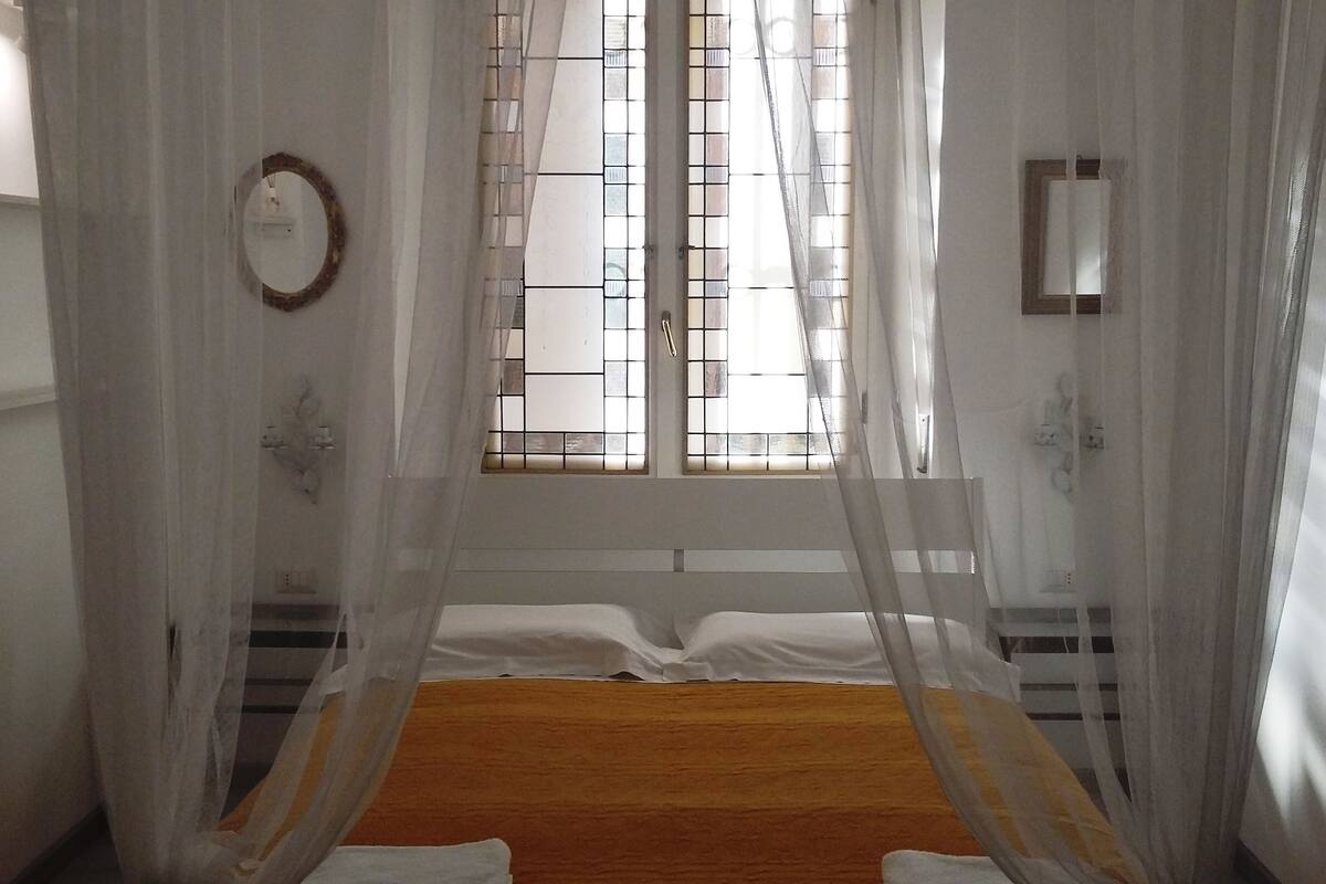 Ein Schlafzimmer mit einem Bett, einem Fenster mit Gitterstäben, einem Spiegel und Vorhängen.