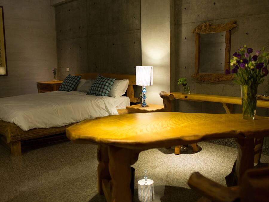 A hotel room with a bed, bedside table, lamp, and a small table with a vase of flowers.