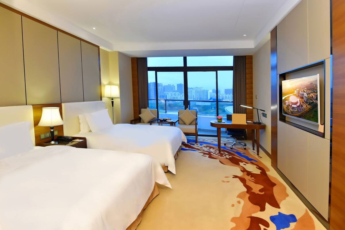 A hotel room with two beds, a desk, a chair, a TV, and a large window with a city view.