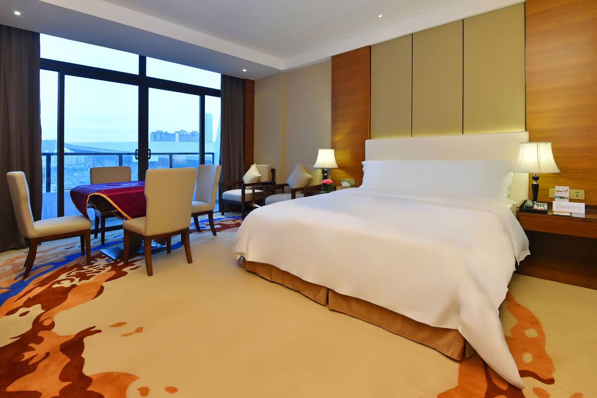 A hotel room with a large bed, two chairs, a dining table, and a view of the city.