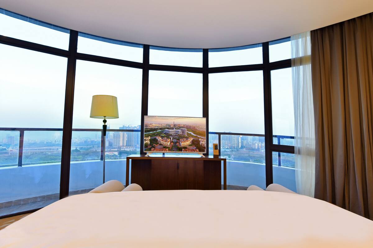 A hotel room with a large bed, a television on a wooden stand, and a view of a cityscape through large windows.