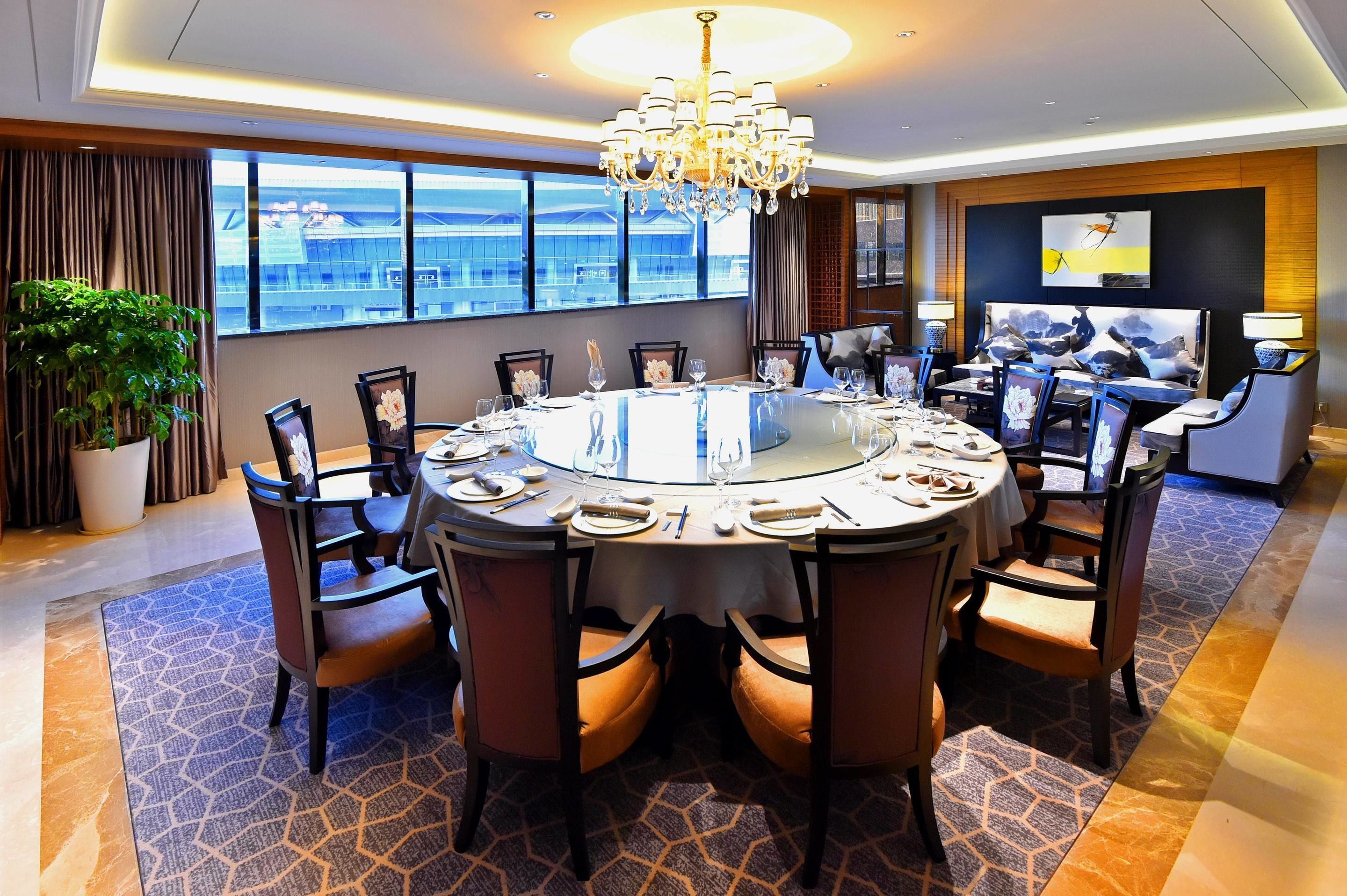 A modern hotel conference room with a round table, comfortable chairs, a chandelier, and large windows offering a city view.