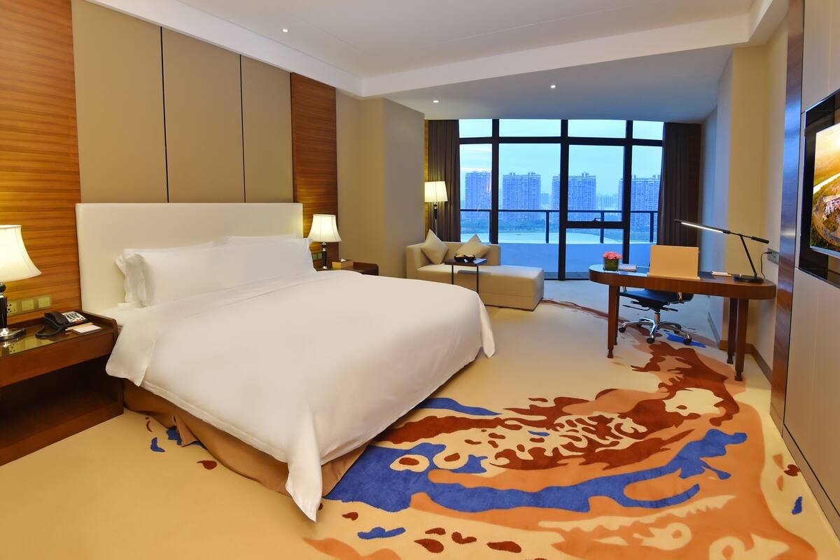 A hotel room with a large bed, a desk, a chair, and a view of the city through large windows.