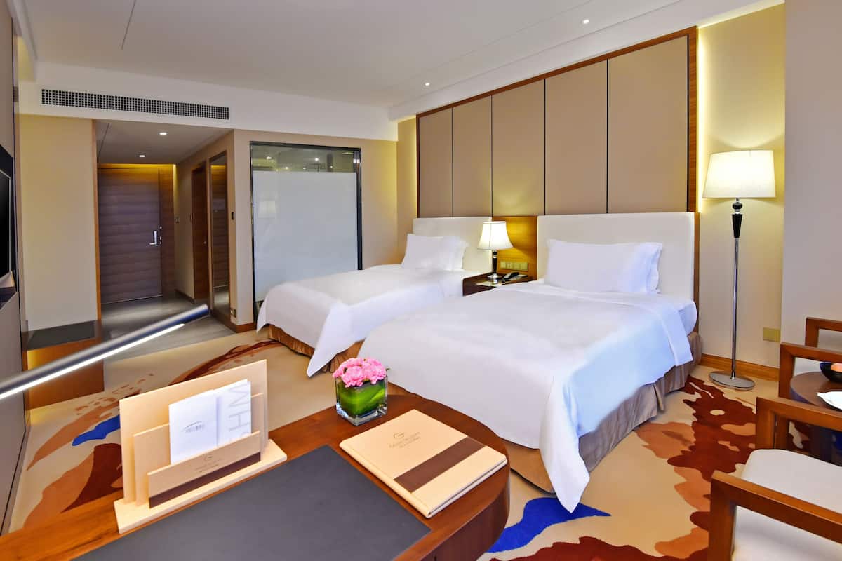 A hotel room with two beds, a desk, a chair, a lamp, a vase with flowers, and a carpet.