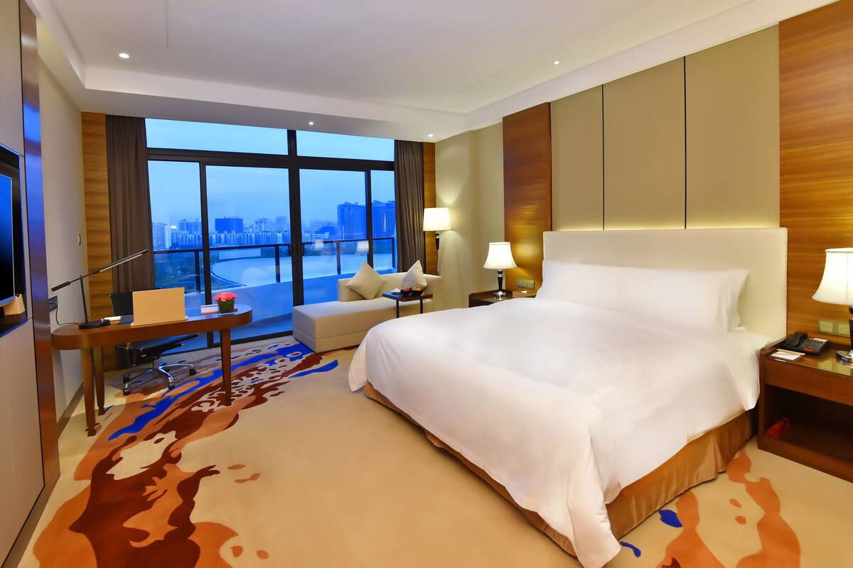 A hotel room with a large bed, a desk with a chair, a sofa, and a view of the cityscape through large windows.