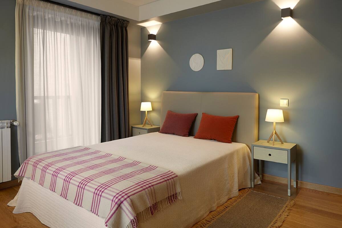 Ein Schlafzimmer mit einem Bett, zwei Nachttischen, einem Fenster mit Vorhängen und Wandleuchten.