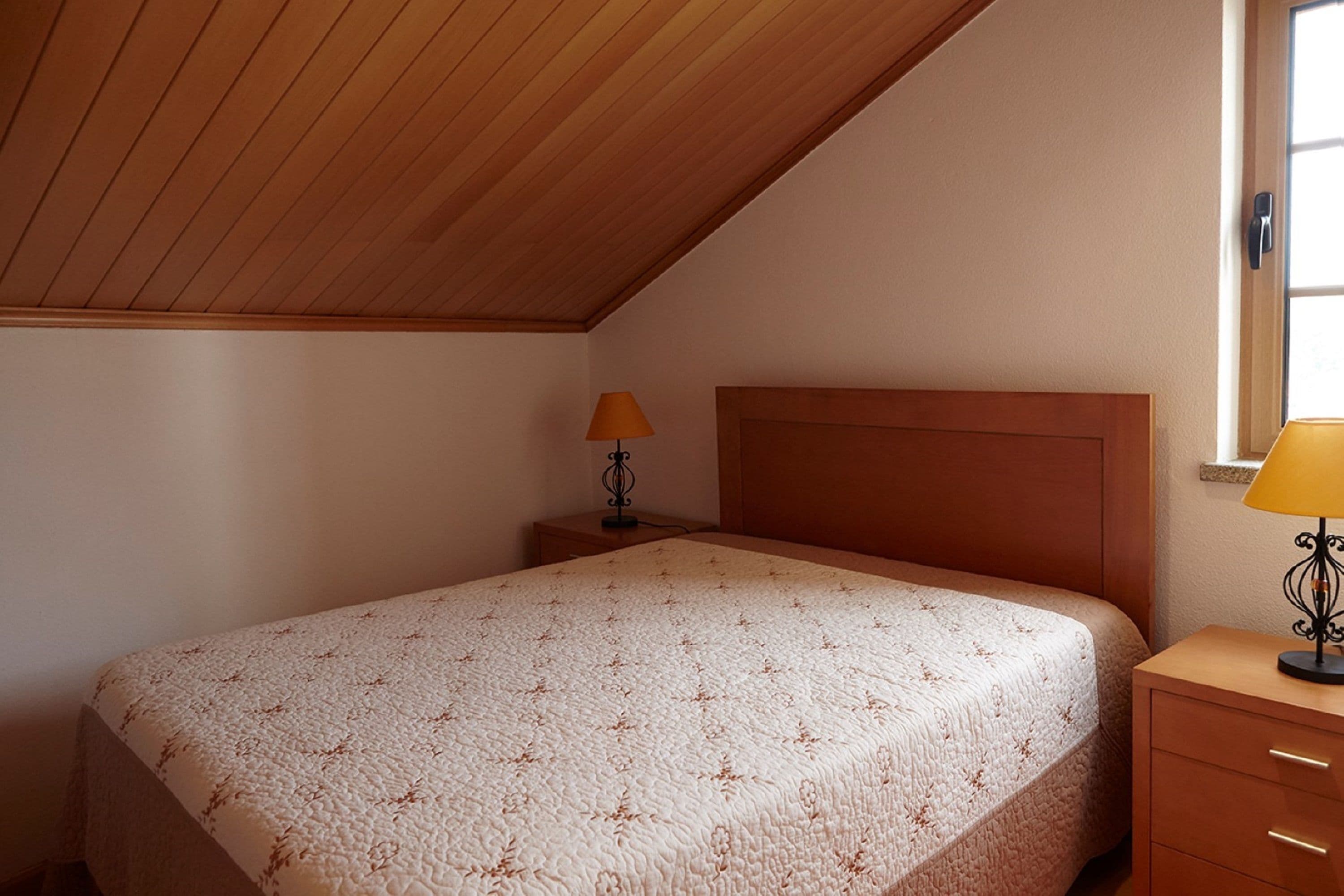 Un dormitorio con techo de madera, una cama con cubrecama estampado, dos mesitas de noche con lámparas y una ventana en la pared derecha.