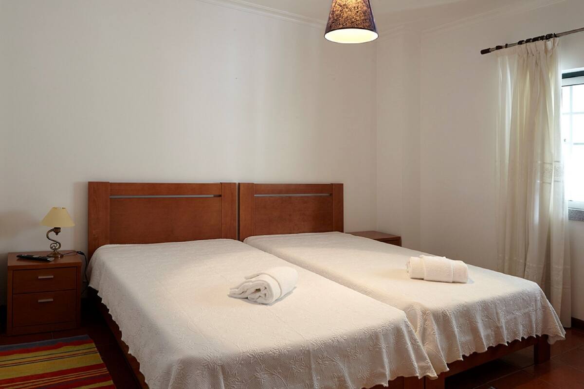 Ein Doppelbett mit Holz Kopfteil, ein Nachttisch mit Lampe und ein Fenster mit Vorhängen.