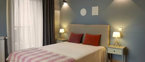 A bedroom with a bed, two bedside tables with lamps, a window with curtains, and a wall with two wall-mounted lights.