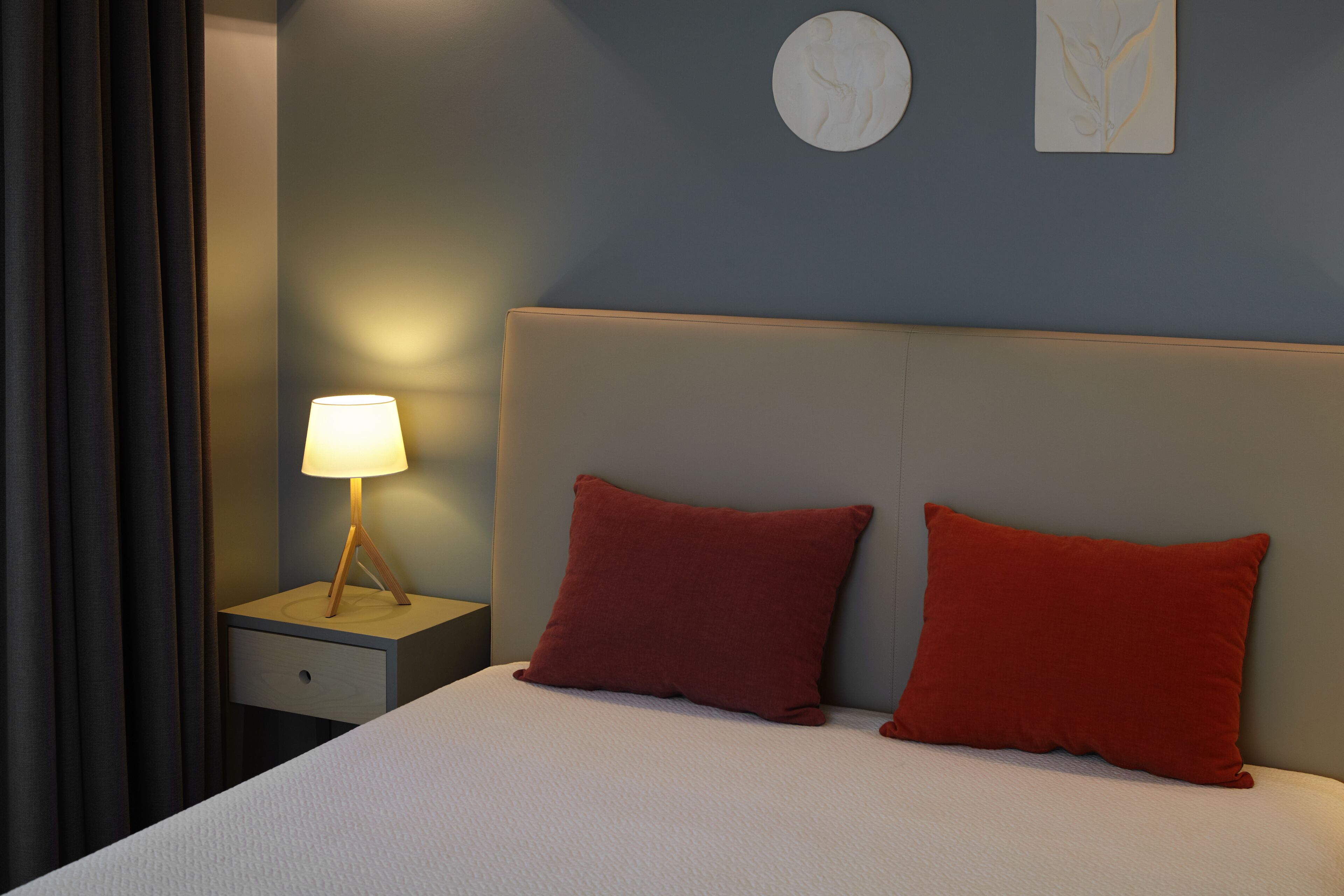 Una cama bien hecha con dos almohadas, una mesita de noche con una lámpara y un reloj de pared.
