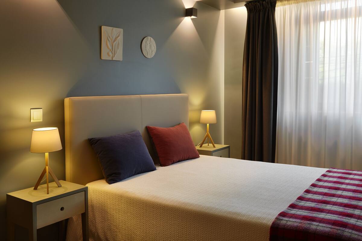 Ein ordentlich bezogenes Bett mit weißem Kopfteil, zwei Nachttischlampen und einem roten und blauen Kopfkissen. An der Wand hängen Uhren und ein Fenster mit transparenten Vorhängen.