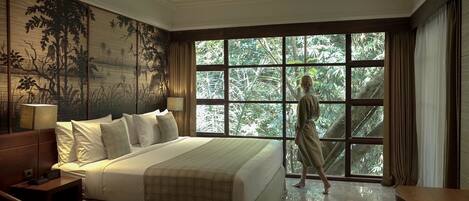 A bedroom with a large bed, a nightstand, a wall-mounted lamp, and a window with a view of greenery.