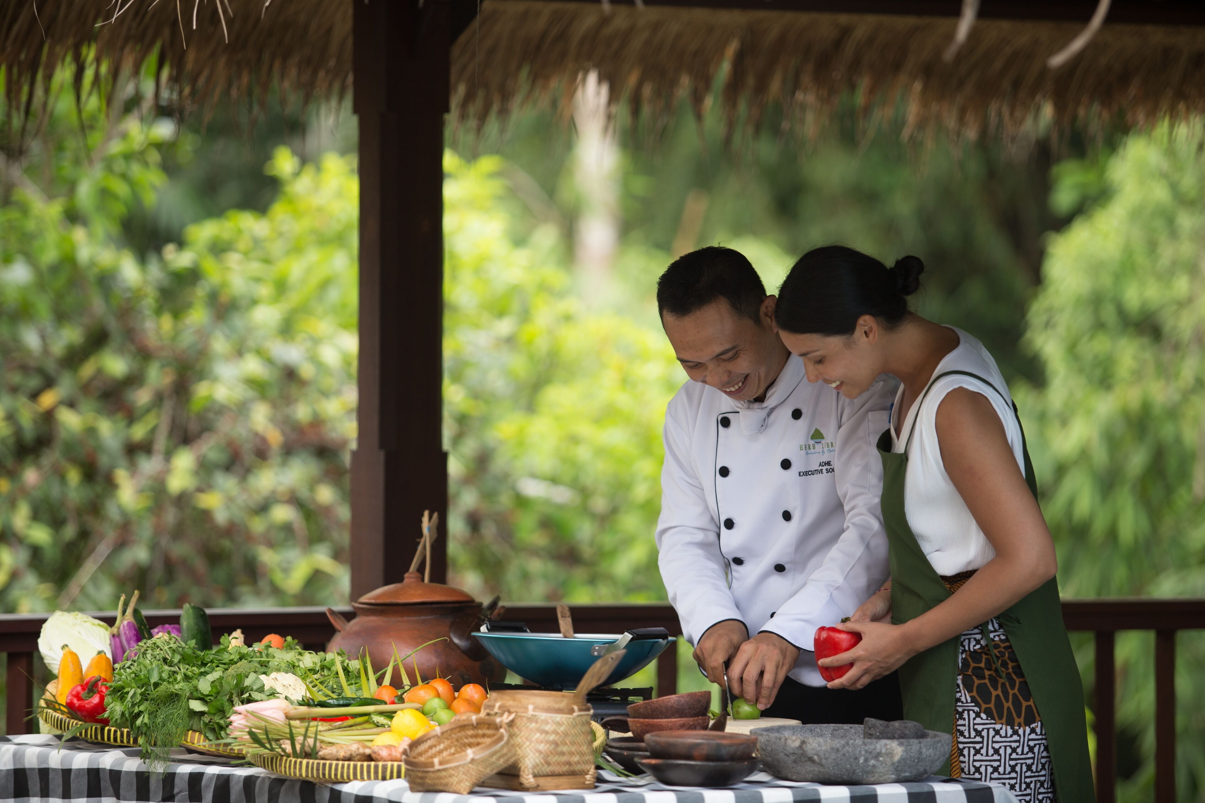 A chef and a woman preparing food outdoors with a table full of vegetables and cooking utensils.