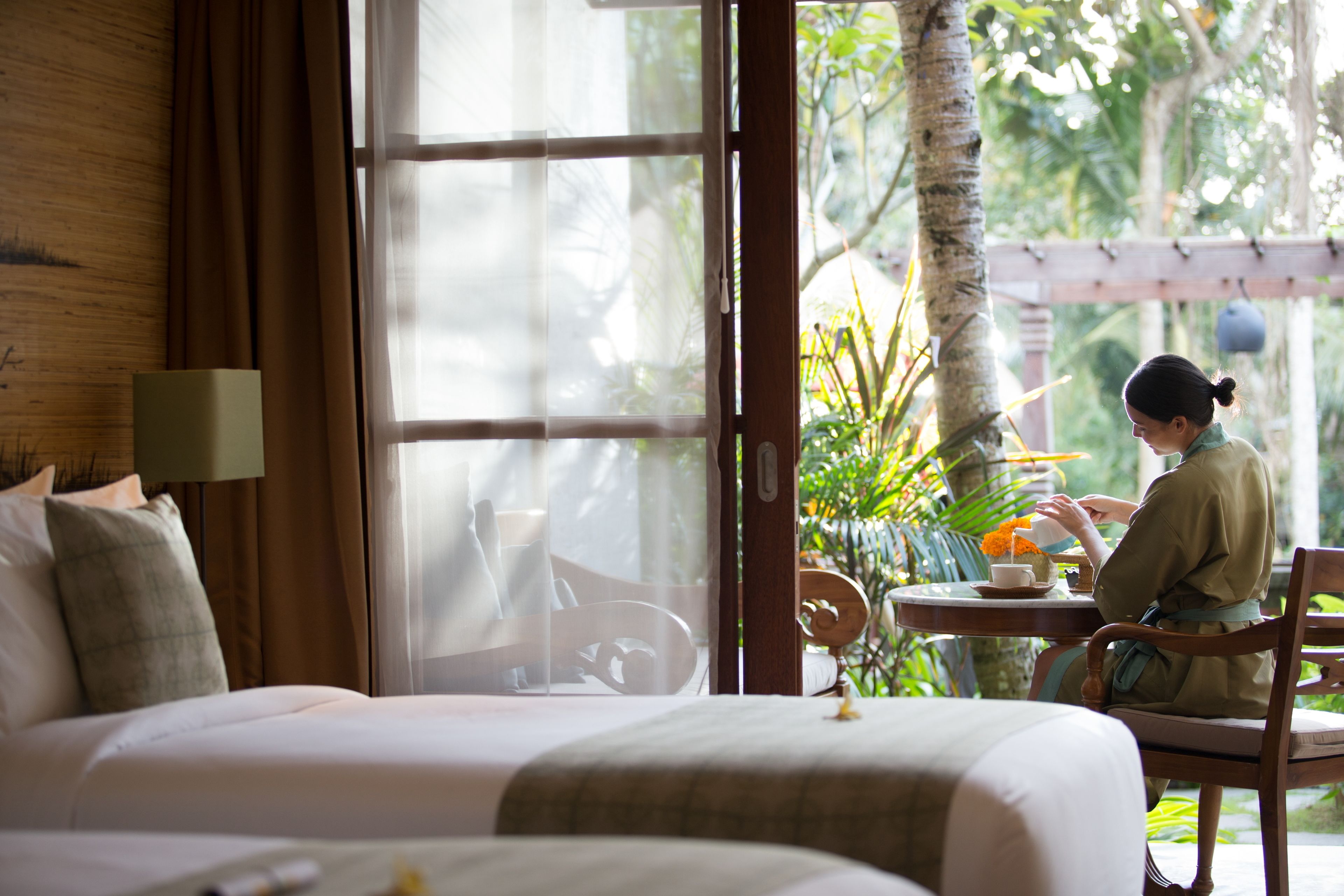 A hotel room with a bed, a table with a cup and saucer, and a view of greenery outside.