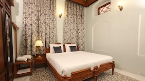 A bedroom with a wooden bed, patterned curtains, a nightstand with a lamp, and a small table with a towel. - Taj Mahal Hotel Abids (Hyderabad)