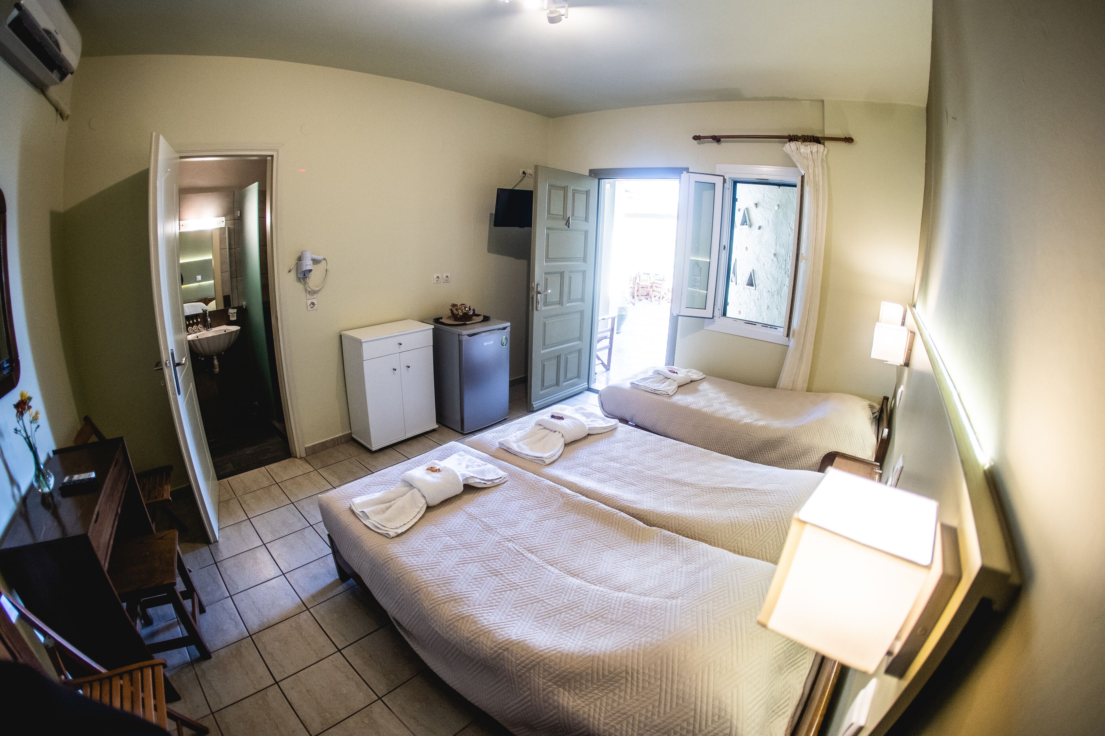Habitación de hotel con dos camas, un escritorio pequeño, una silla, una lámpara de escritorio, una ventana con cortinas, un televisor y un refrigerador miniatura.