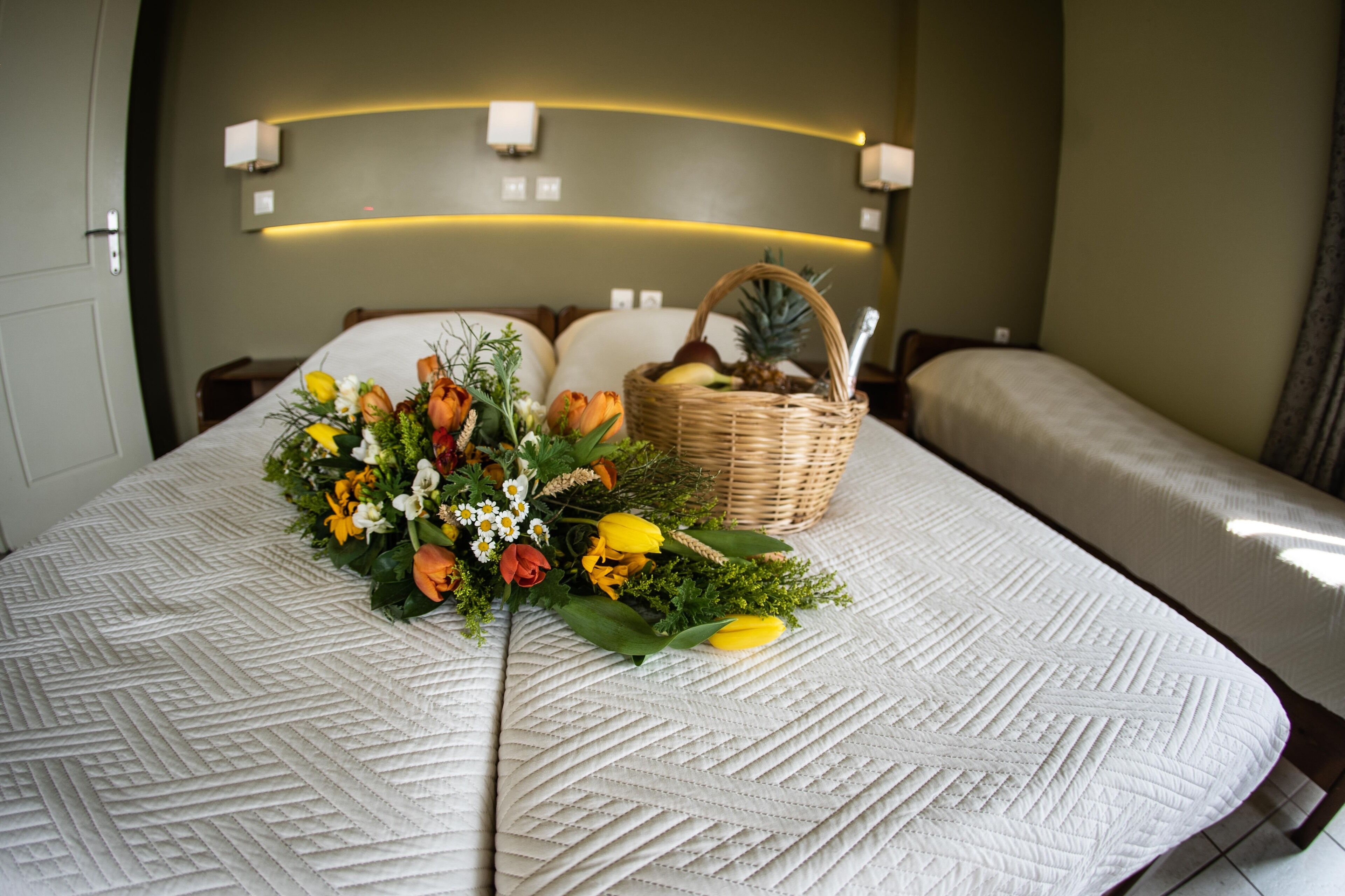 Una cama bien hecha con colcha blanca, un arreglo floral sobre la cama y una cesta de mimbre con frutas.