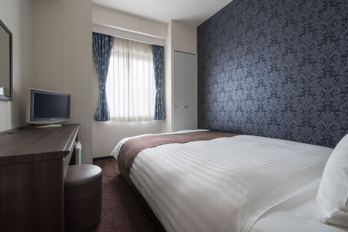Habitación de hotel con cama, escritorio con televisor, armario y ventana con cortinas.