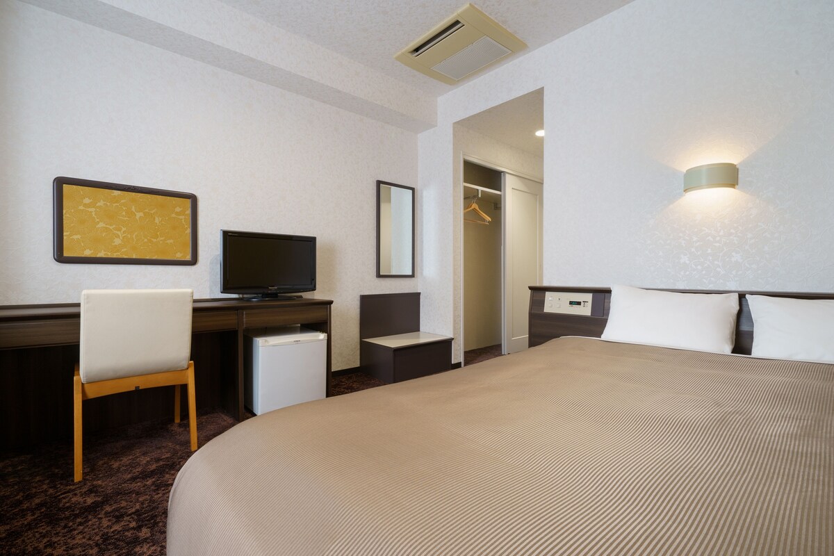 Habitación de hotel con una cama grande, un escritorio con silla, un televisor, un armario y una lámpara de pared.