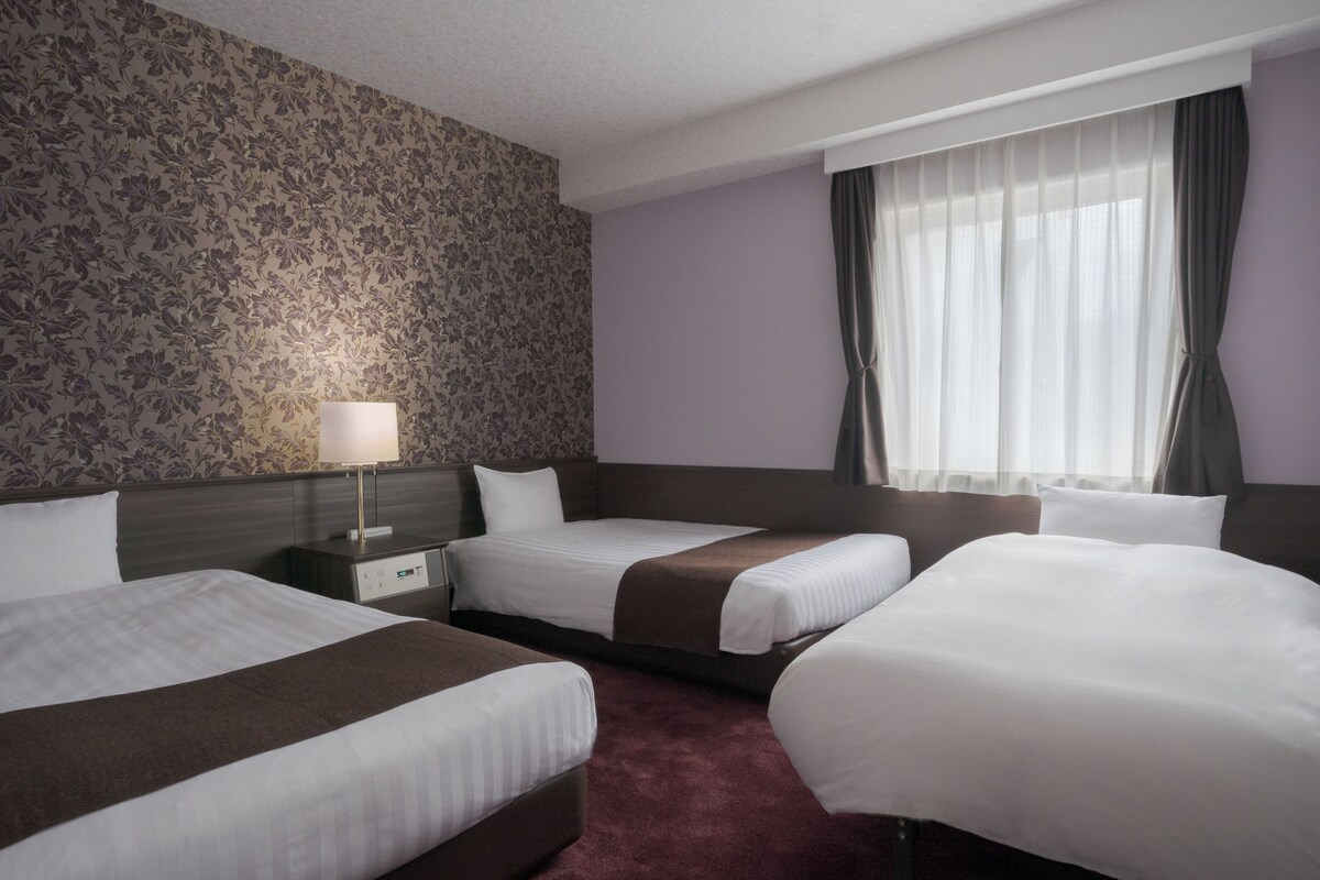 Habitación de hotel con dos camas, una mesita de noche, una lámpara y una ventana con cortinas.