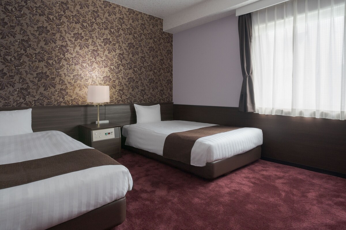 Una habitación de hotel con dos camas, una mesita de noche, una lámpara y una ventana con cortinas.
