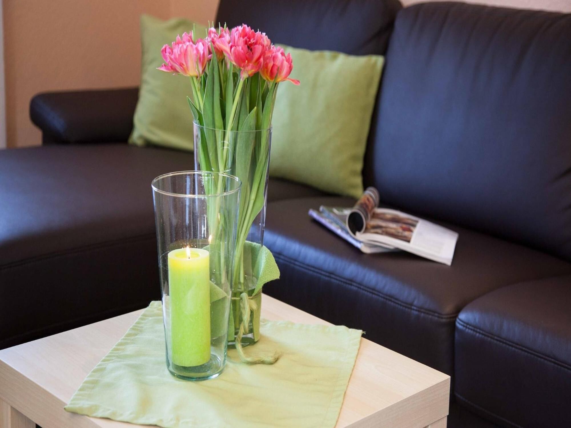 Un vase en verre contenant des fleurs roses et une bougie allumée, posé sur une table devant un canapé en cuir noir agrémenté de coussins verts.