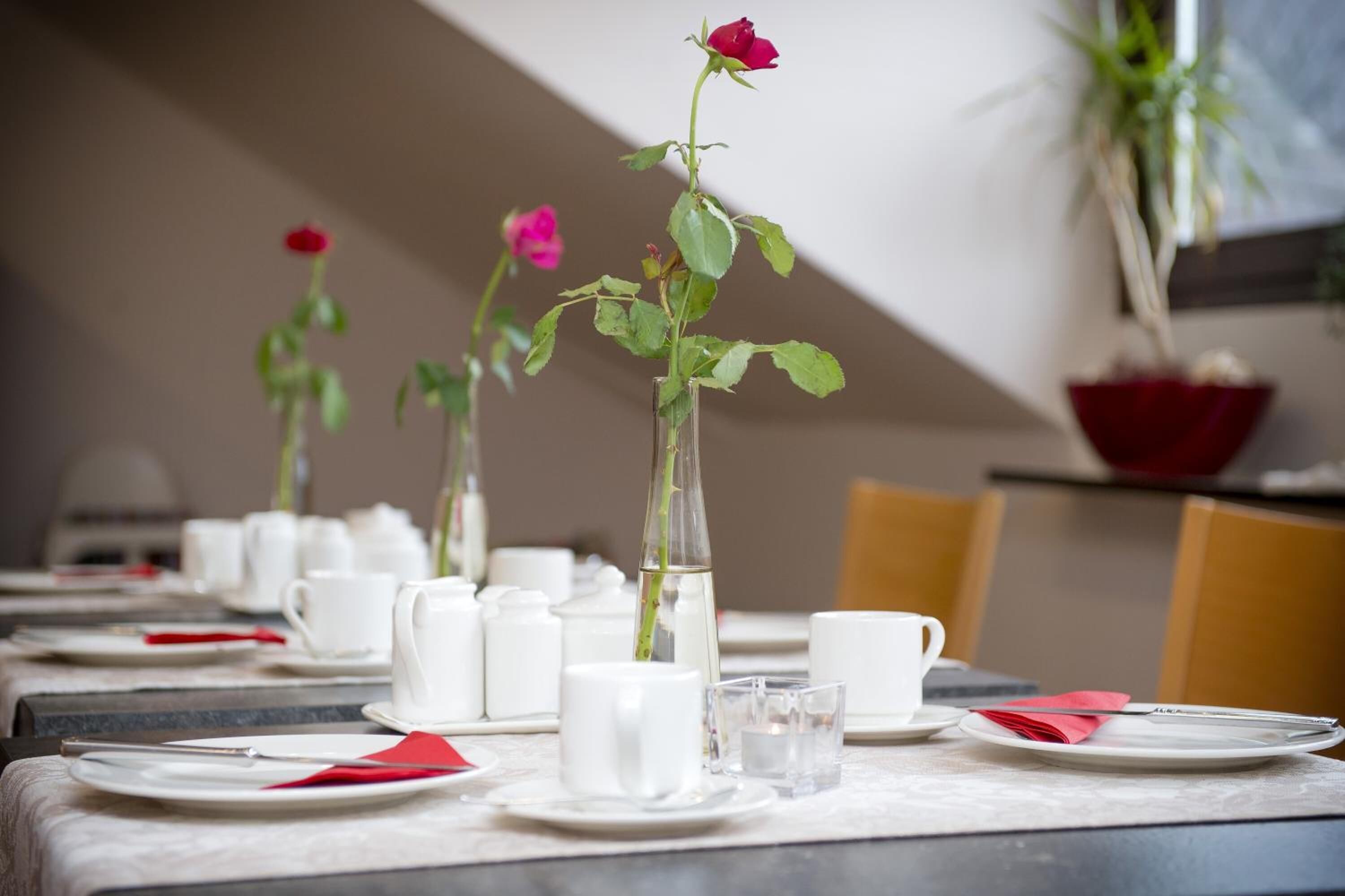 A dining table set with white plates, cups, and a red napkin, with a rose in a clear vase.
