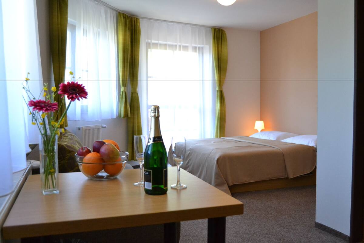 A hotel room with a bed, a bedside table with a bottle of champagne, a bowl of fruit, and a vase with flowers.