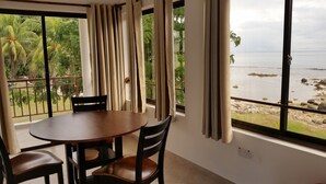 Una habitación con una mesa redonda y dos sillas, una ventana con cortinas y vistas a una costa rocosa con palmeras.