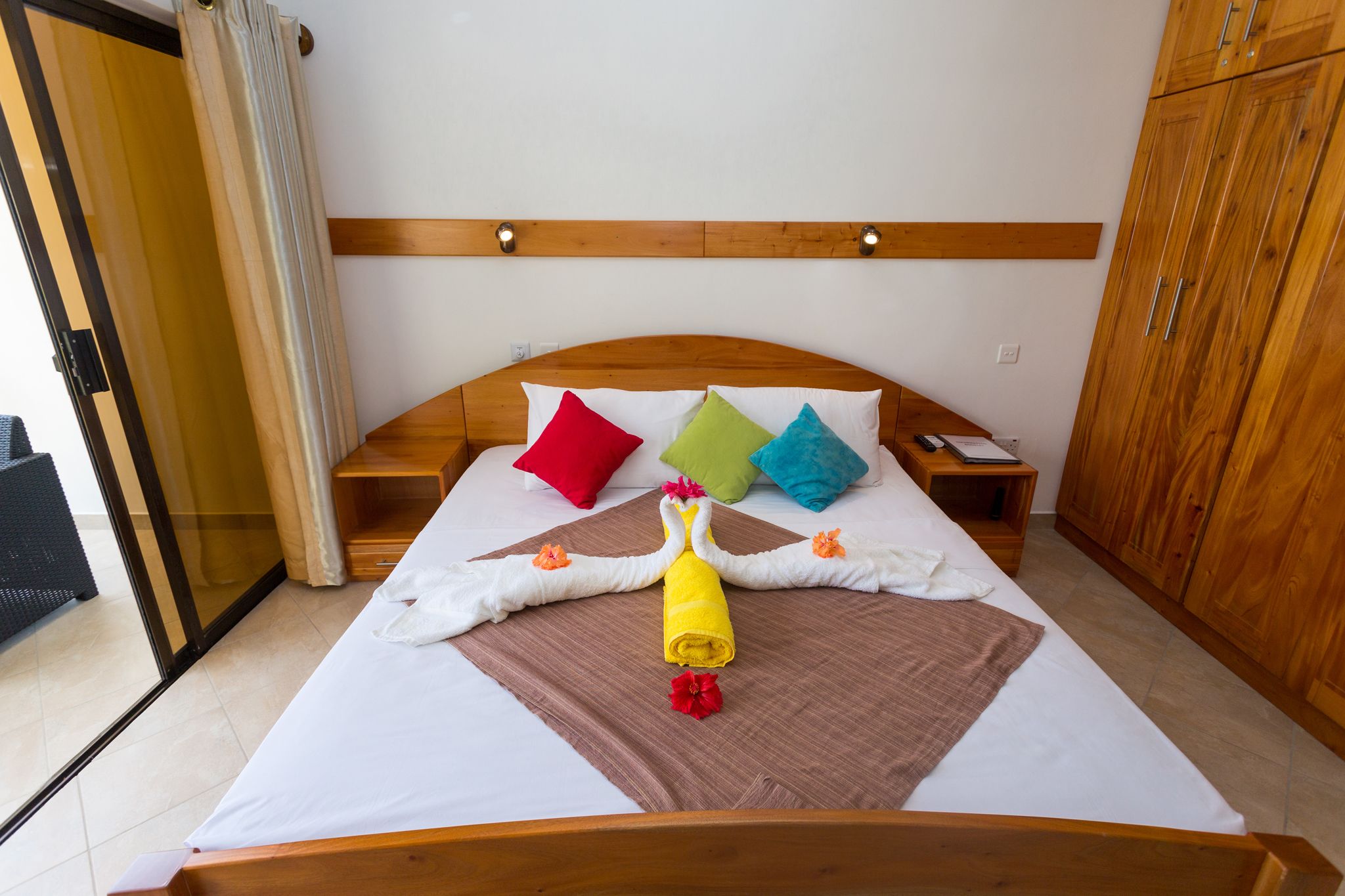 Hotelkamer met een bed, houten hoofdeinde, kleurrijke kussens, handdoekenset en houten kasten.