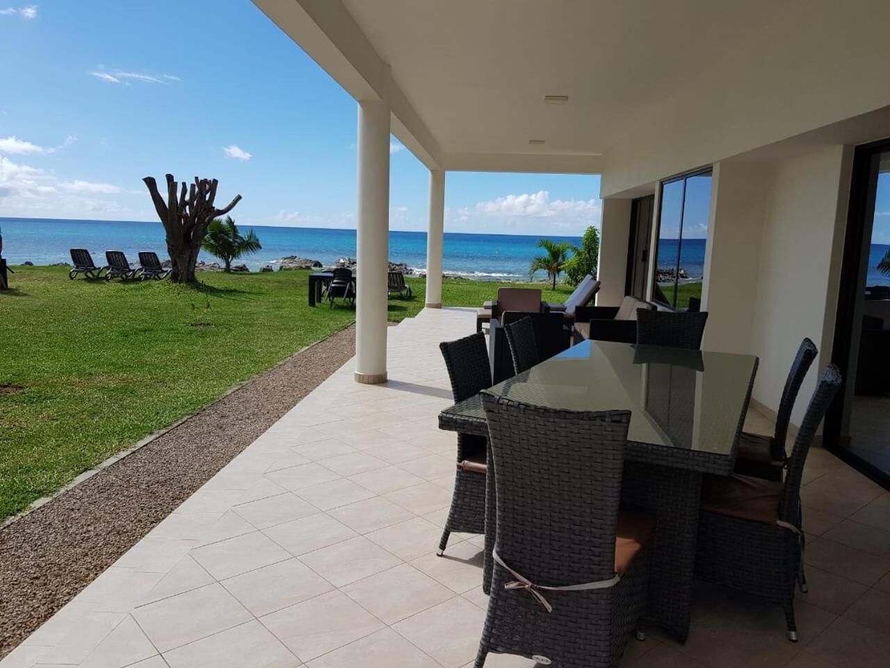 Een terras buiten met een eettafel en stoelen, met uitzicht op een strand met ligbedden en een palmboom.